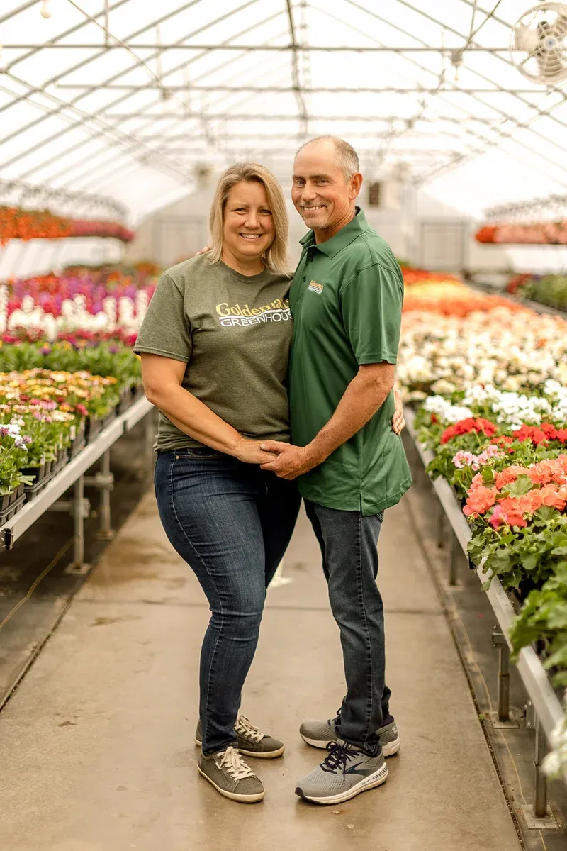 Alan and Kris Goldeman standing together in a greenhouse full of blooming flowers